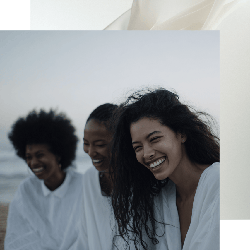 Three women laughing together on a beach with ocean in the background